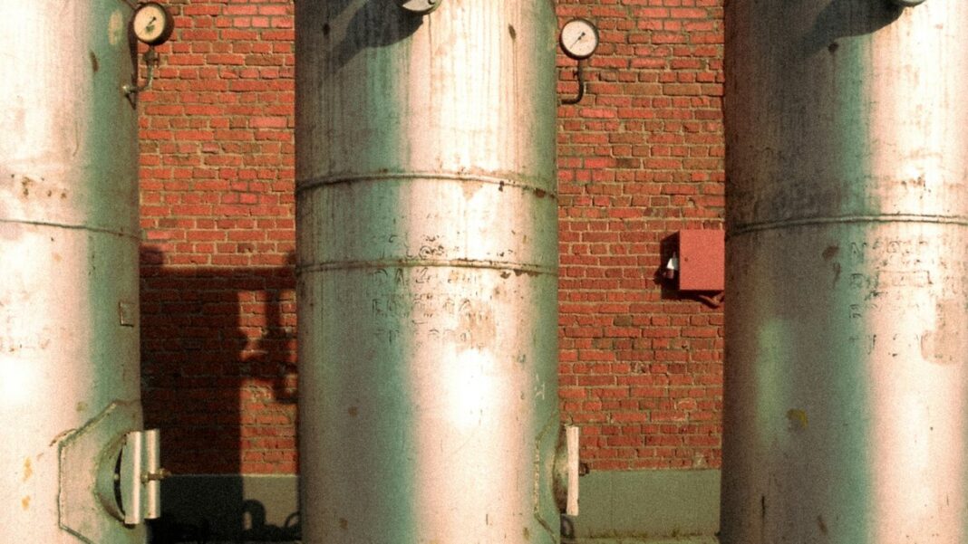 Drei hohe Metallspeicher mit Manometern vor einer roten Ziegelwand